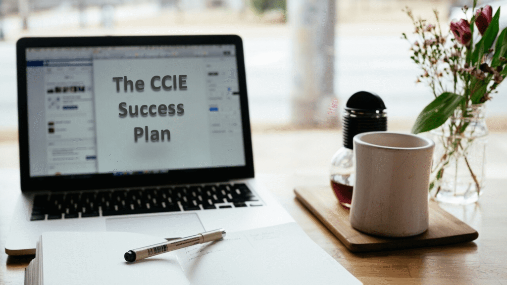 The CCIE Success Strategy Plan A laptop displaying the text “The CCIE Success Plan” sits on a wooden table next to an open notebook, a pen, a white coffee mug on a saucer, and a small vase with flowers. The scene is set near a window with a blurred view outside.